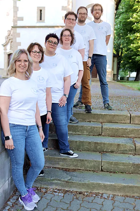 7 Mitarbeiter frontal fotografiert in weißen T-Shirts auf einer Treppe